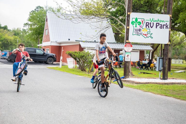 Two boys ride bicycles, one performing a wheelie, near the entrance of Vinton RV Park. In the background, families are gathered at picnic tables under a tree, and a large sign reading “Vinton RV Park” is visible next to a red building with a metal roof.