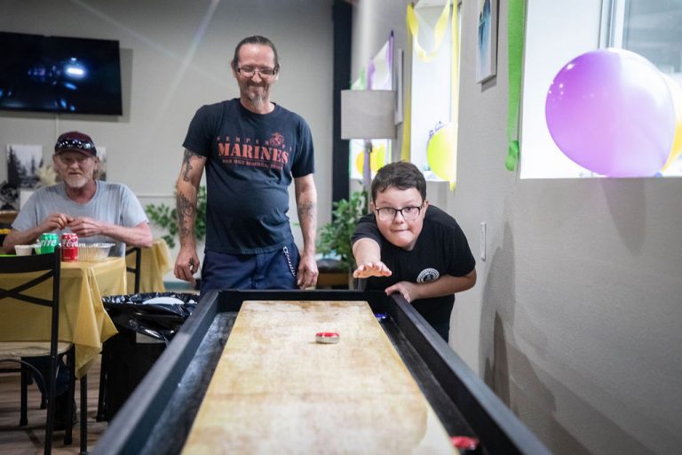 A young boy concentrates as he plays shuffleboard inside a recreation room at Vinton RV Park, with two men watching and smiling in the background. The room is decorated with colorful balloons and streamers, creating a festive atmosphere.