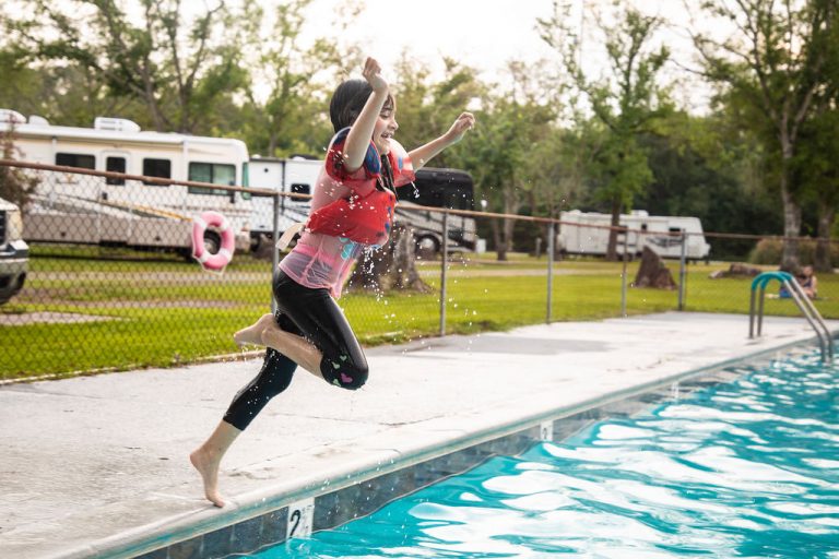A young girl wearing a red life vest jumps joyfully into a swimming pool at Vinton RV Park. Behind her, several RVs are parked on the grassy grounds, and a chain-link fence lines the pool area. Trees and greenery surround the park on a bright day.